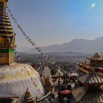 A photo of Swayambhu taken from a nearby roof shows the main stupa and temple complex in the foreground and Kathmandu in the background.