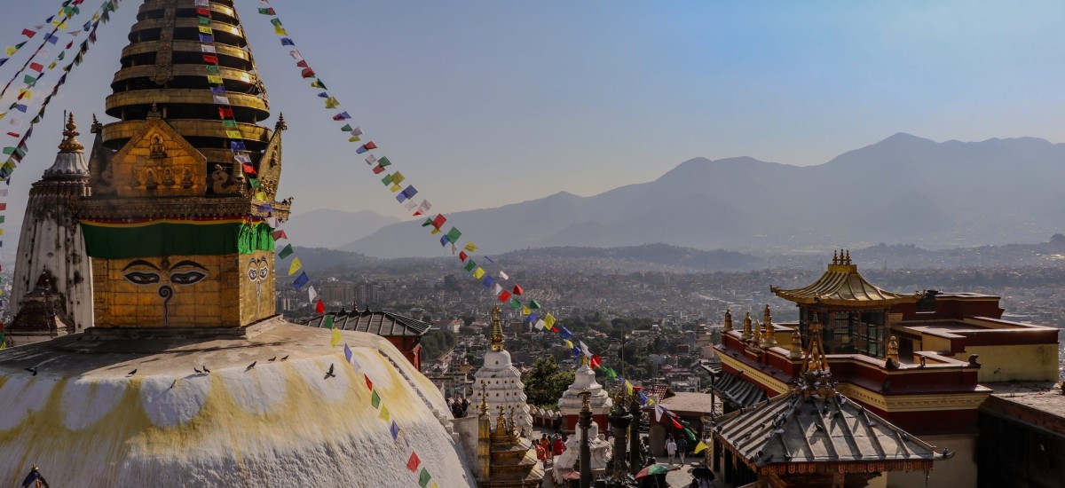 A photo of Swayambhu taken from a nearby roof shows the main stupa and temple complex in the foreground and Kathmandu in the background.